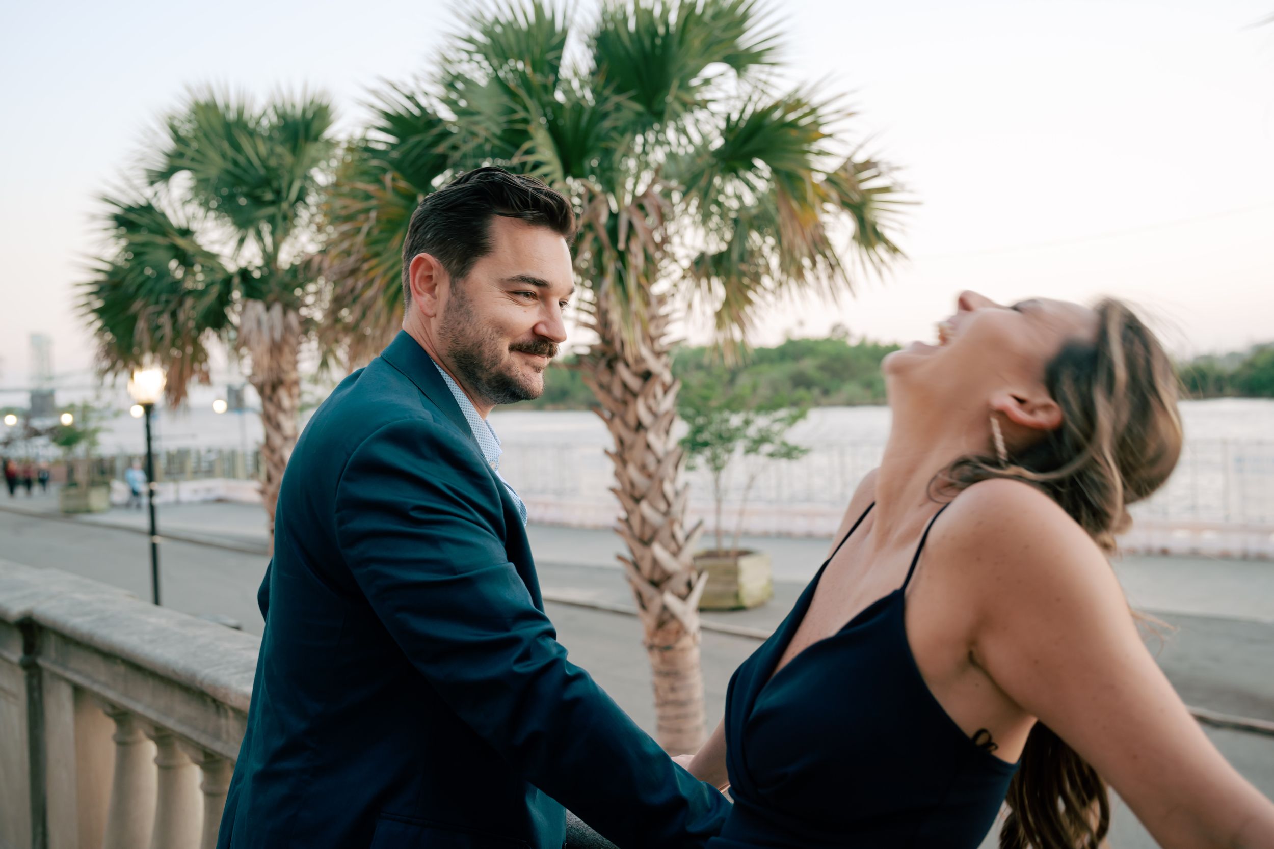 A couple shares a joyful moment outdoors near palm trees and a waterfront, dressed in elegant navy attire.