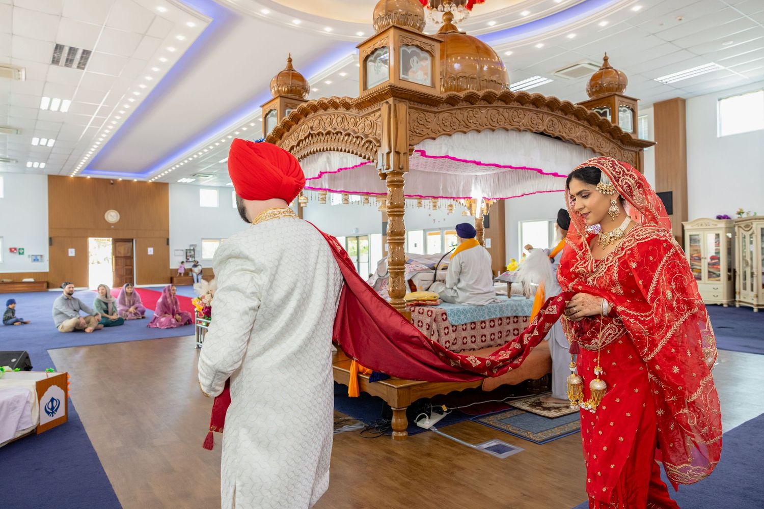 Sikh bride and groom in traditional red and white attire during a wedding ceremony inside a Gurdwara.