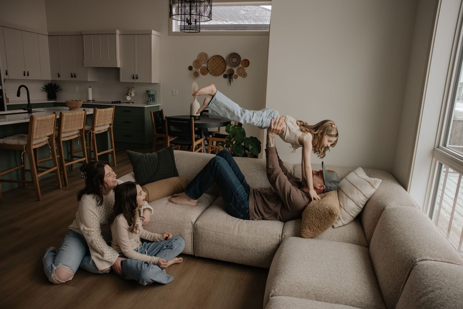 Family playing on a cozy beige sectional sofa in a modern open-plan living room, with a child being lifted in the air.