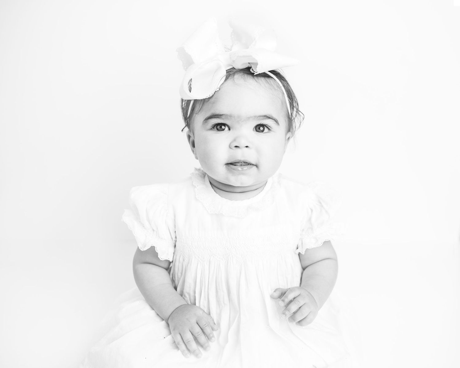 Black and white portrait of adorable baby girl in white dress and bow headband looking at camera.