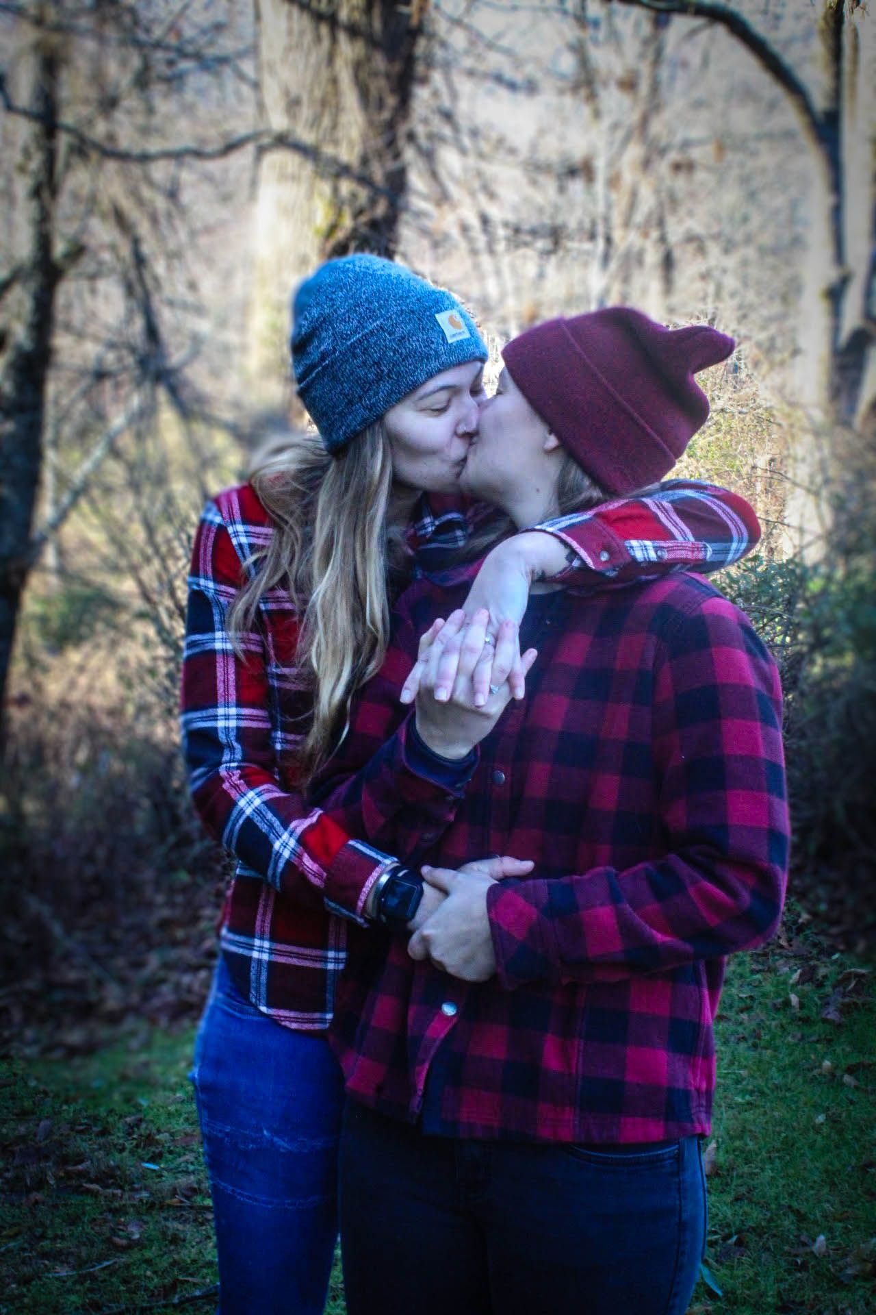 Two people in winter clothing and beanies share a romantic kiss in a woodsy setting.