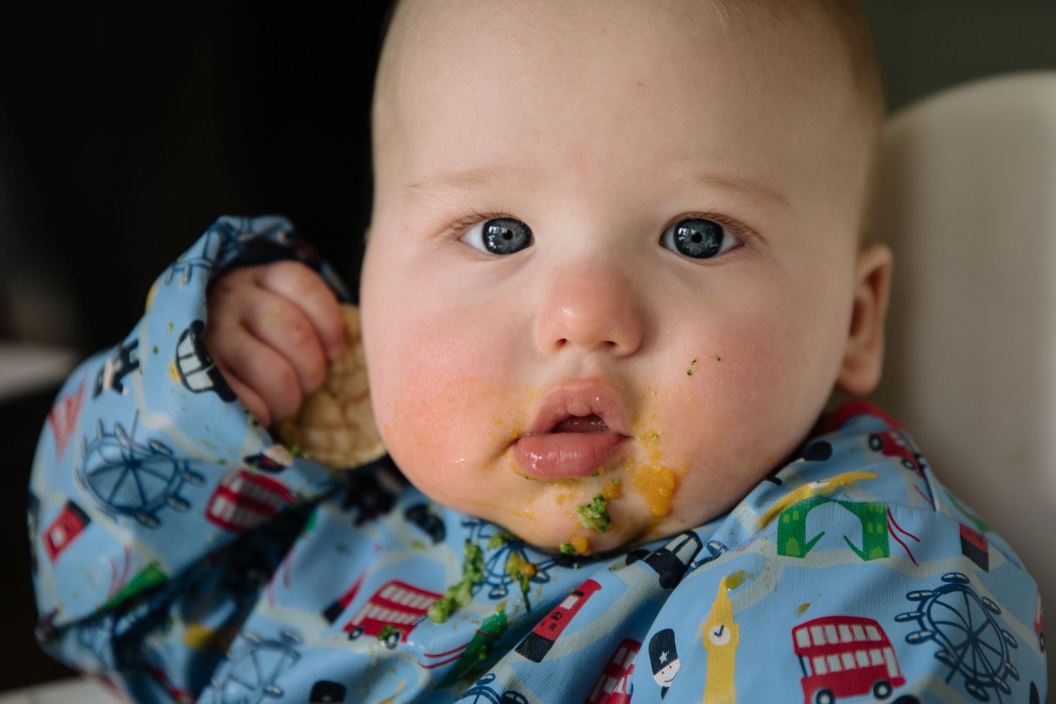 Baby in dinosaur print bib has food smeared around mouth while eating.