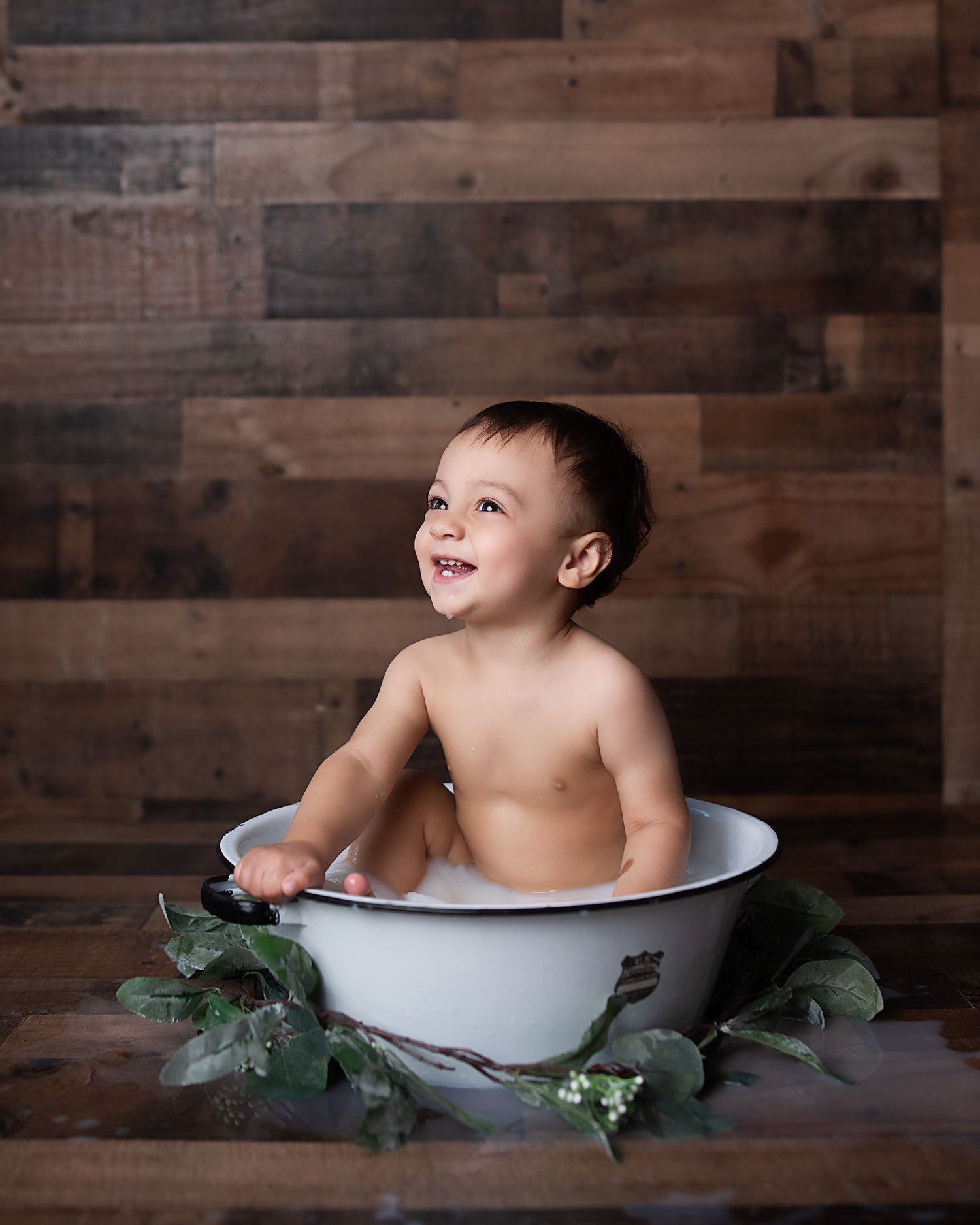 Smiling baby boy sitting in vintage white enamel tub with greenery during milestone bath photo session.