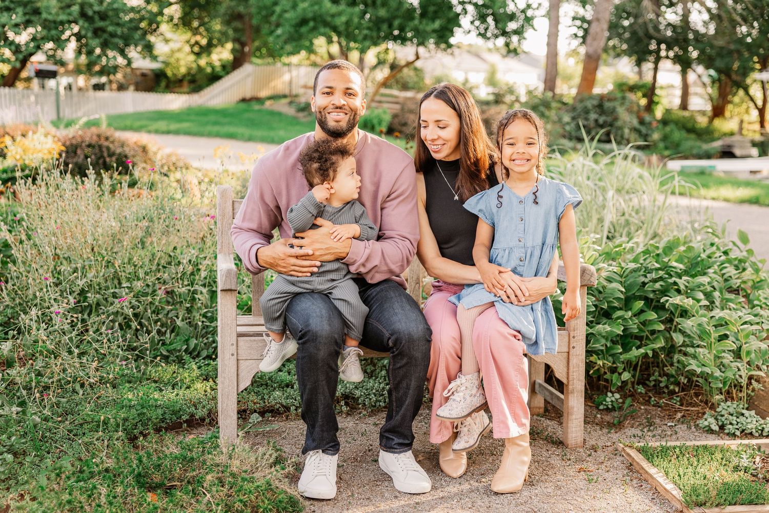 Family sits together on a wooden bench in a lush garden setting.