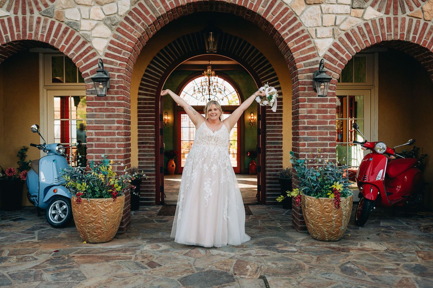 A wedding dress displayed in an ornate brick archway entrance with vintage scooters on either side.