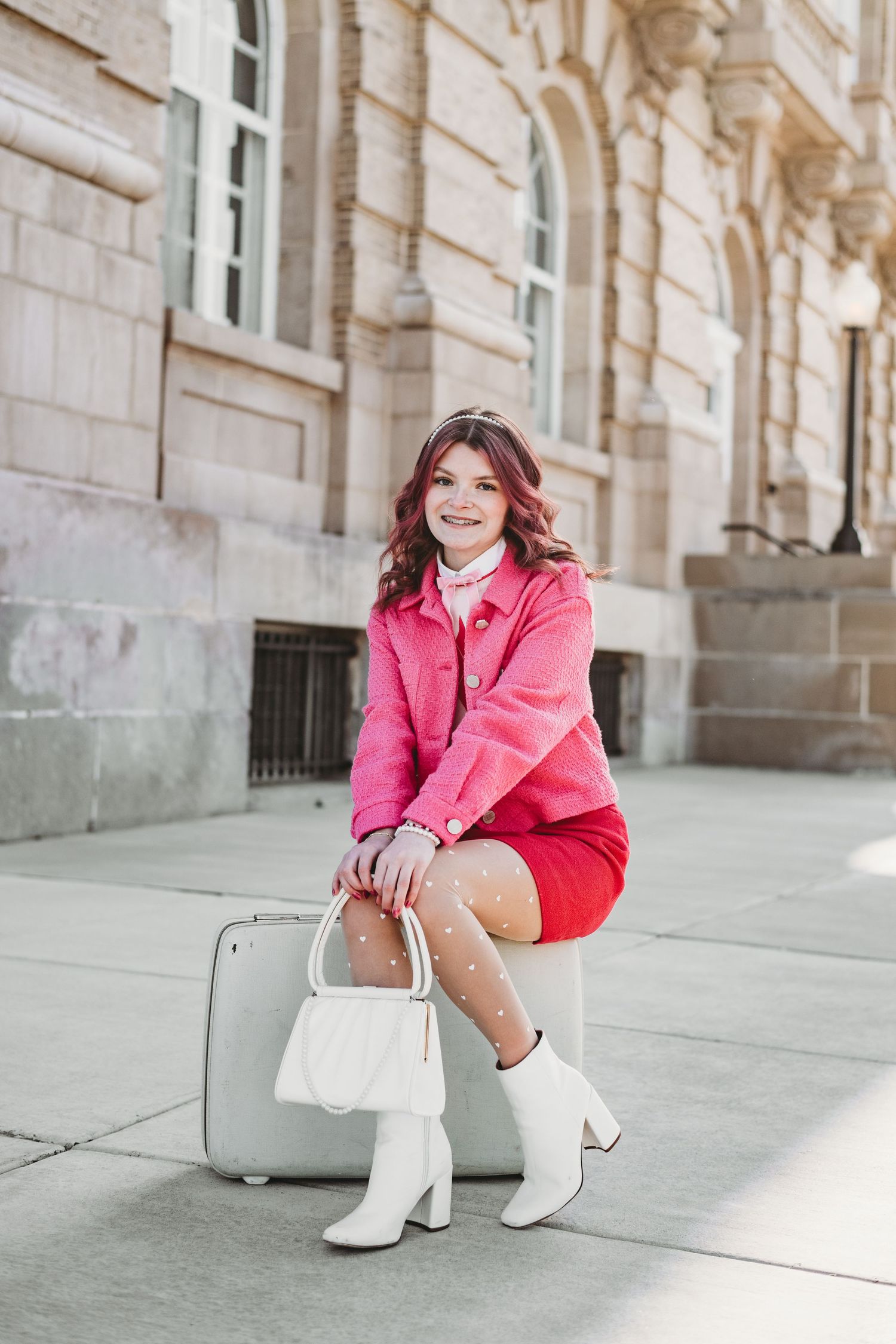 Fashion portrait in pink jacket and white boots sitting on stone steps of historic building.
