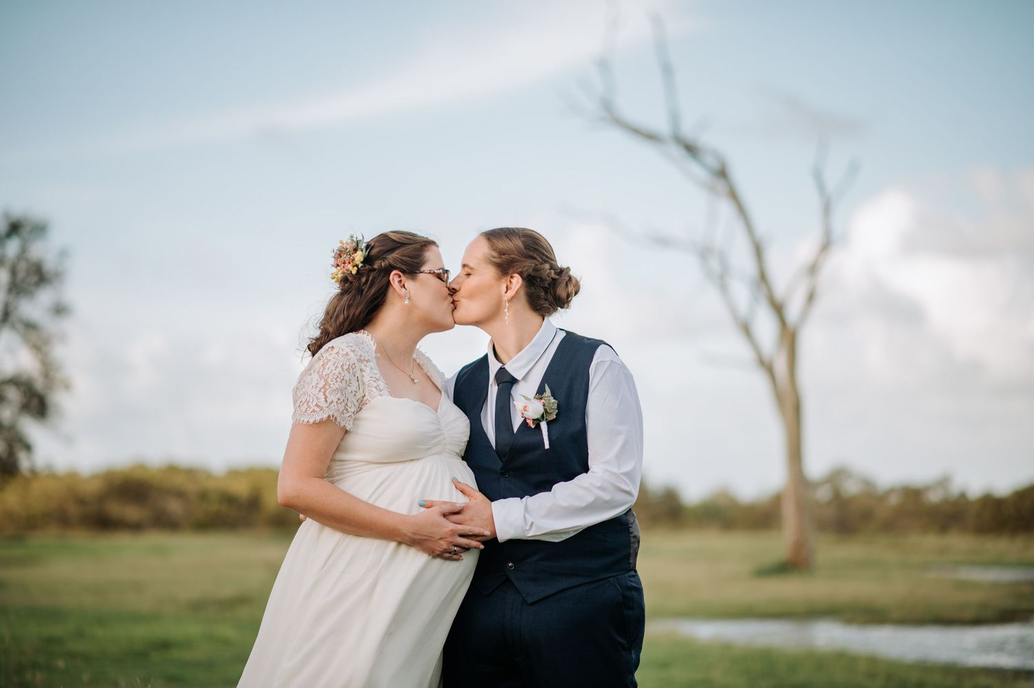 Couple shares intimate moment in outdoor setting with bare tree in distance.