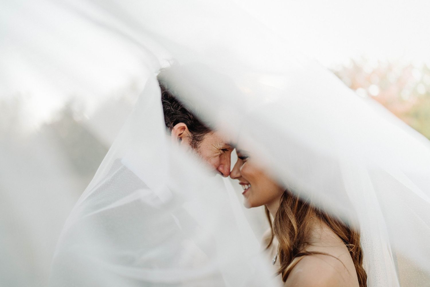 Playful couple shares laughter under billowing white veil.
