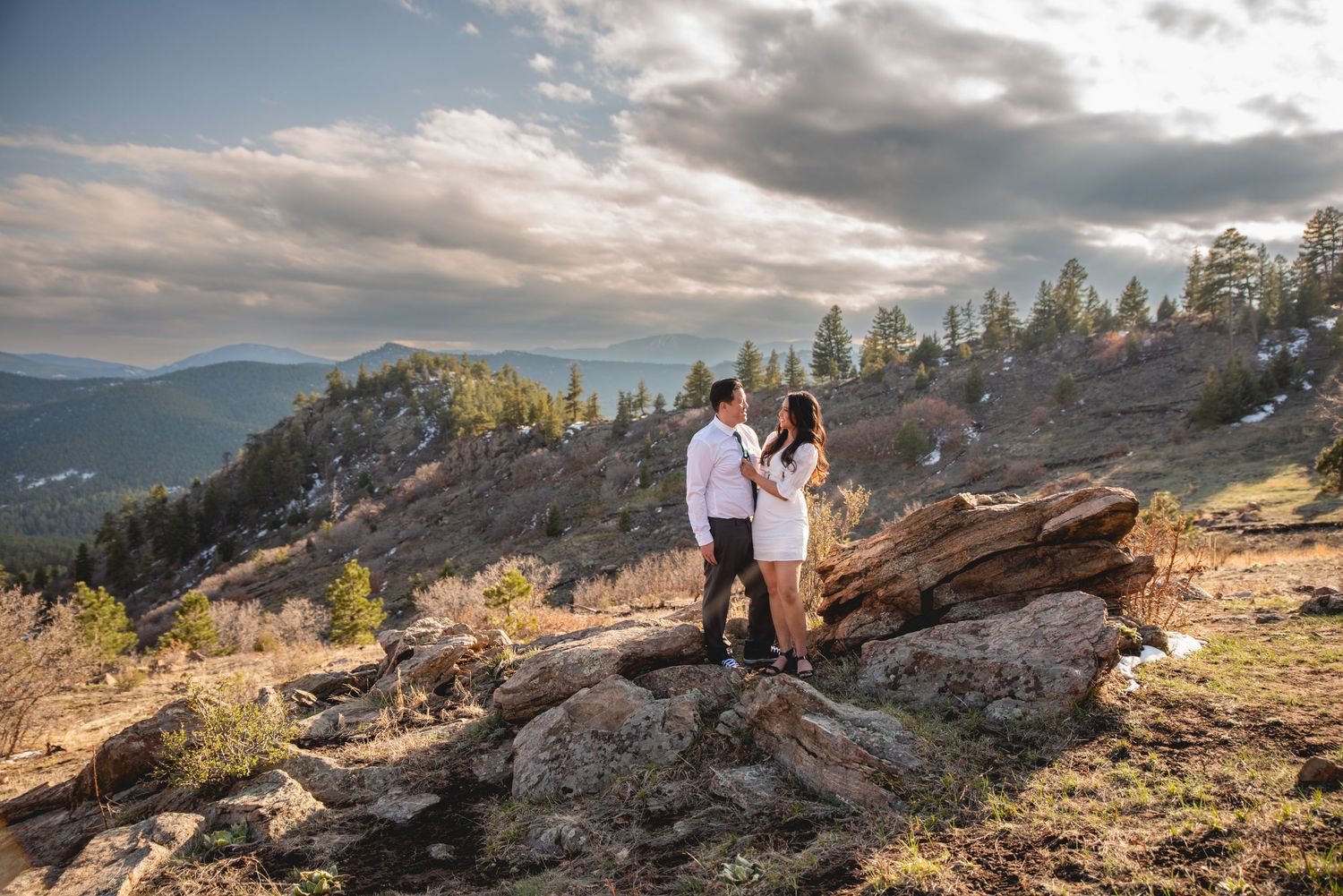 A couple stands together on a rocky mountain overlook during golden hour with sweeping valley views.