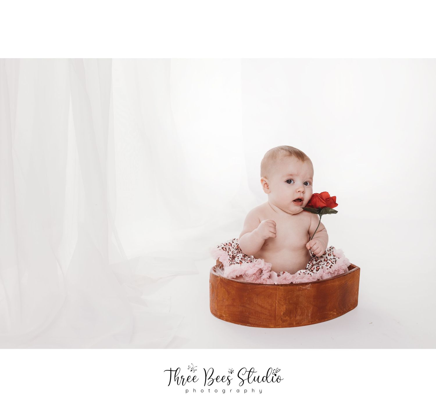 Baby sitting on brown cushion with red flower in minimalist studio setting.