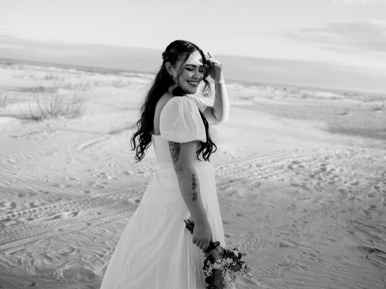 A bride in a flowing white dress poses in a desert landscape holding a bouquet.