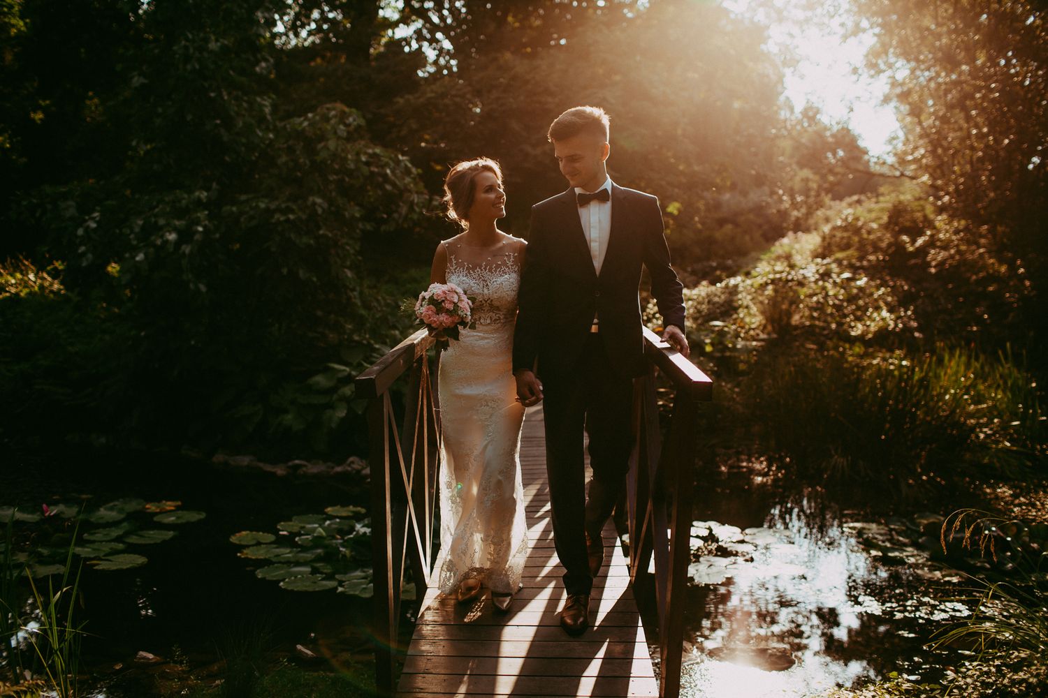 Bride and groom walk hand in hand over a wooden bridge beside a pond at sunset.