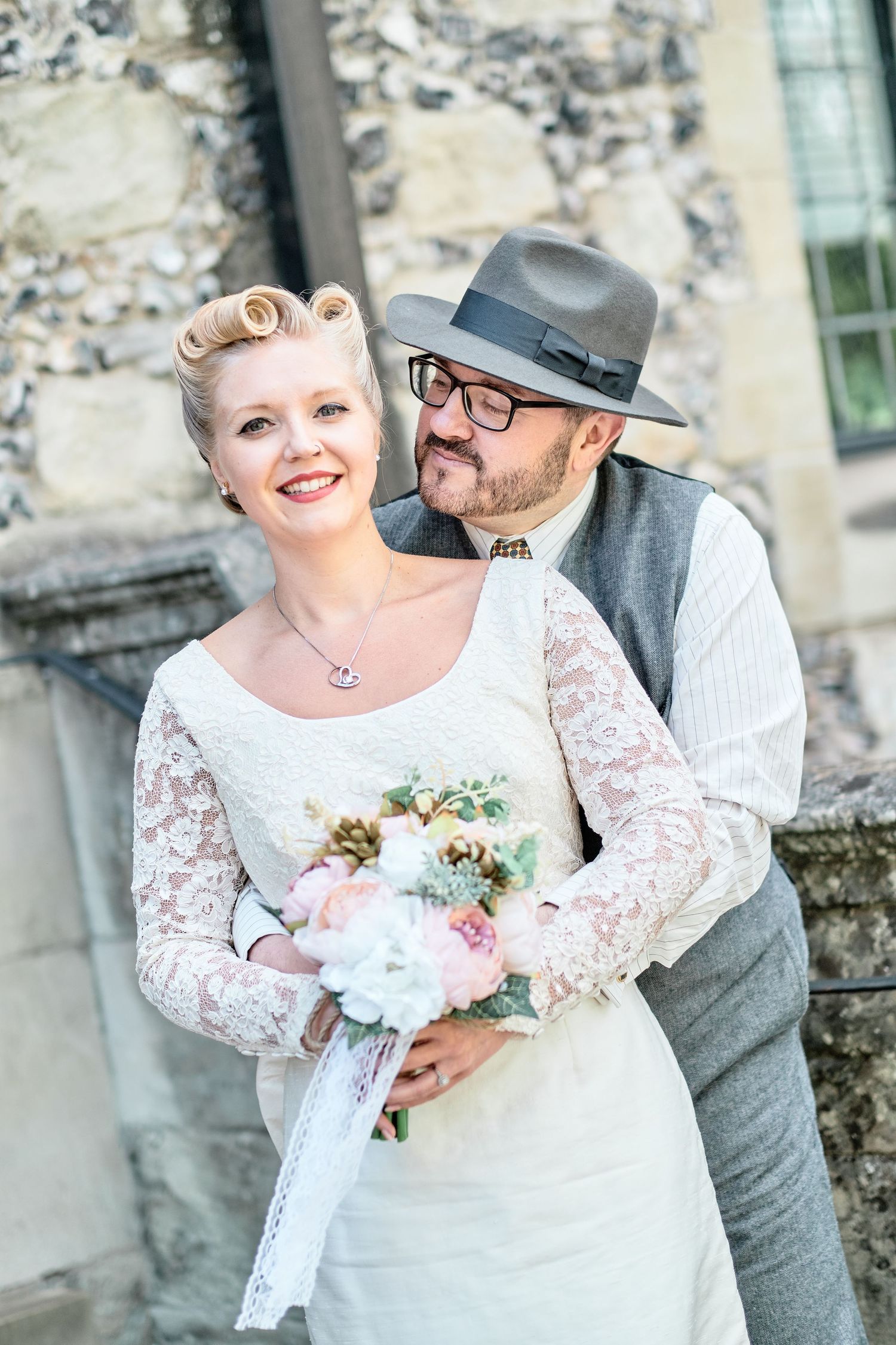 Vintage-styled wedding portrait against stone wall with bride in lace dress.