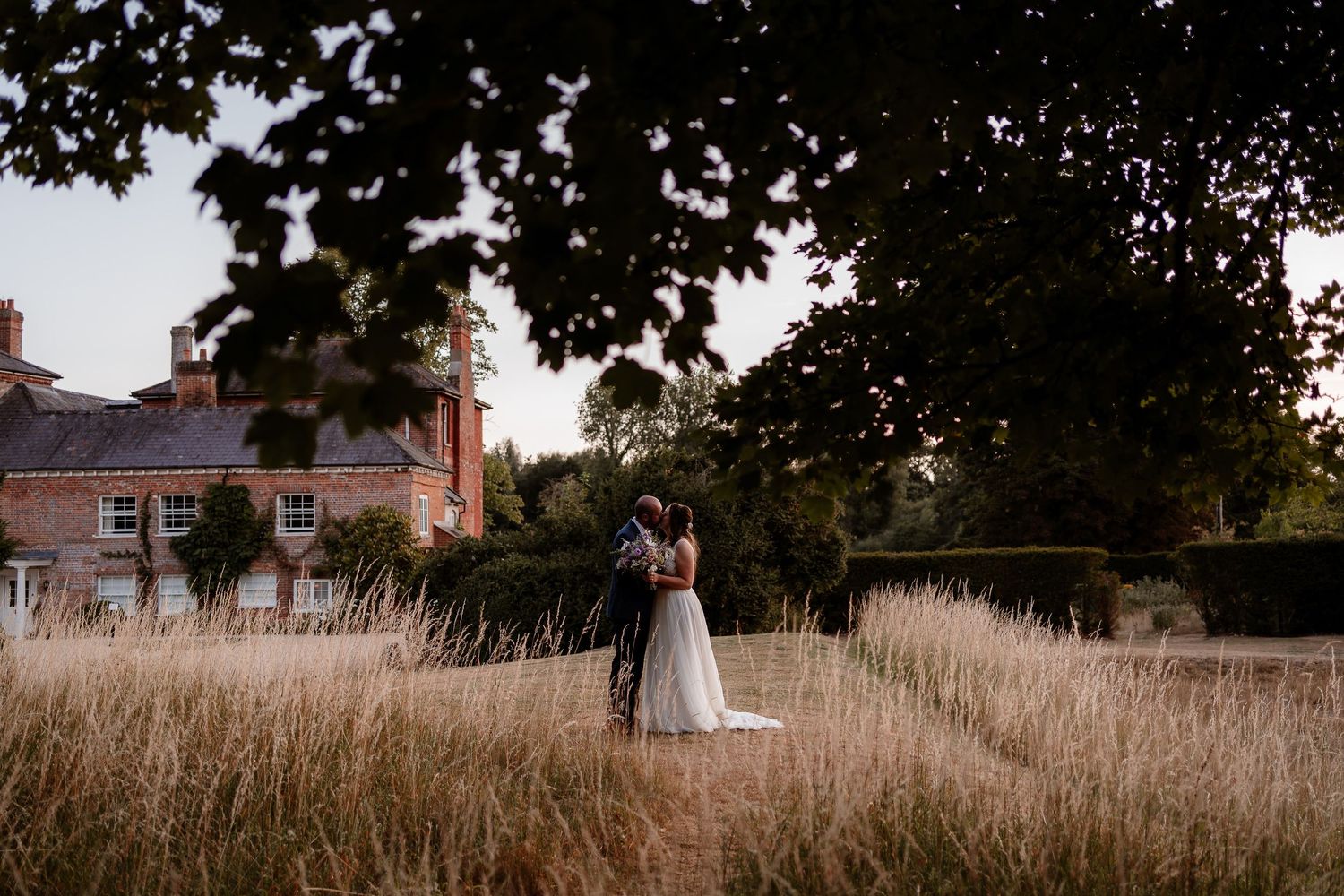A newlywed couple kiss at golden hour among the long grasses at Syrencot wedding venue