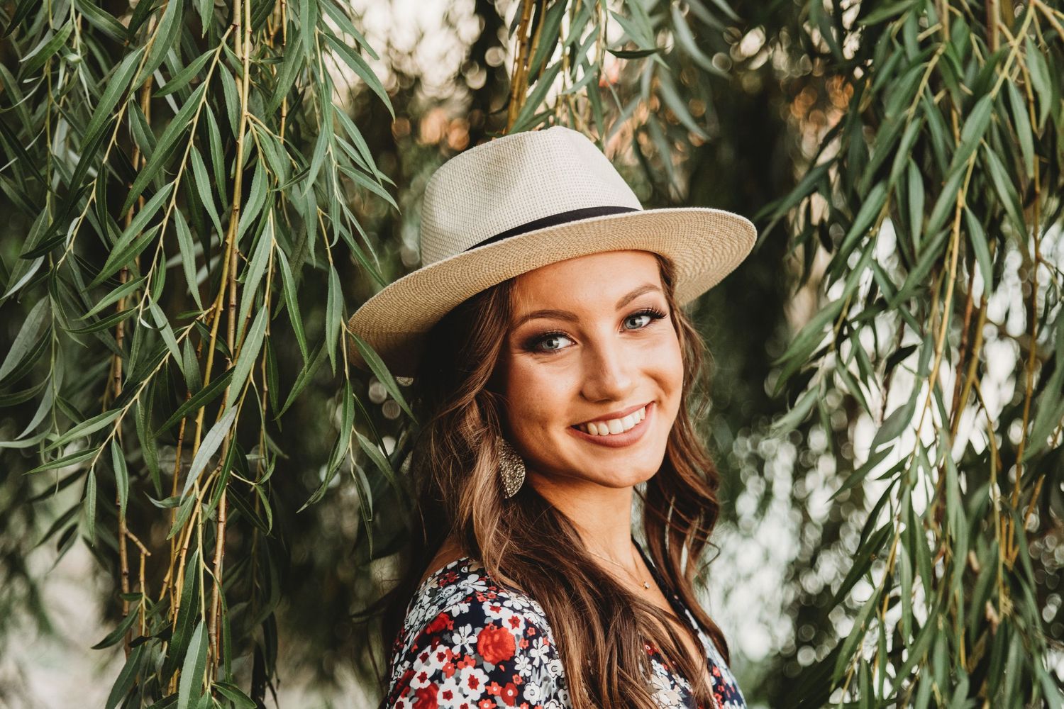 Woman in straw hat and floral dress poses under willow tree branches.