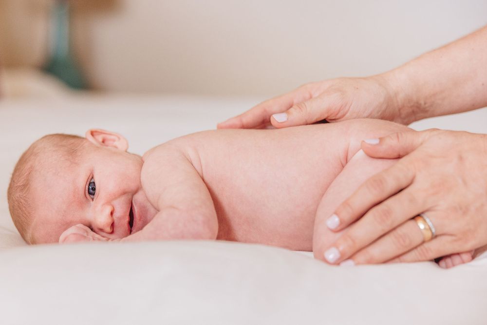 Gentle hands massage a smiling baby during an infant massage session on white bedding.