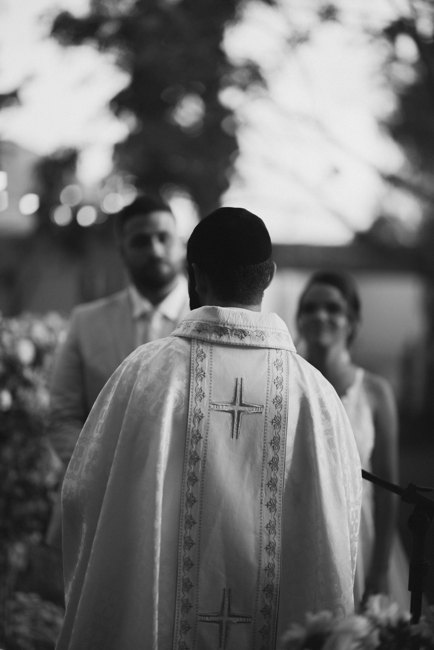 Black and white photo of priest in embroidered robe during outdoor wedding ceremony.