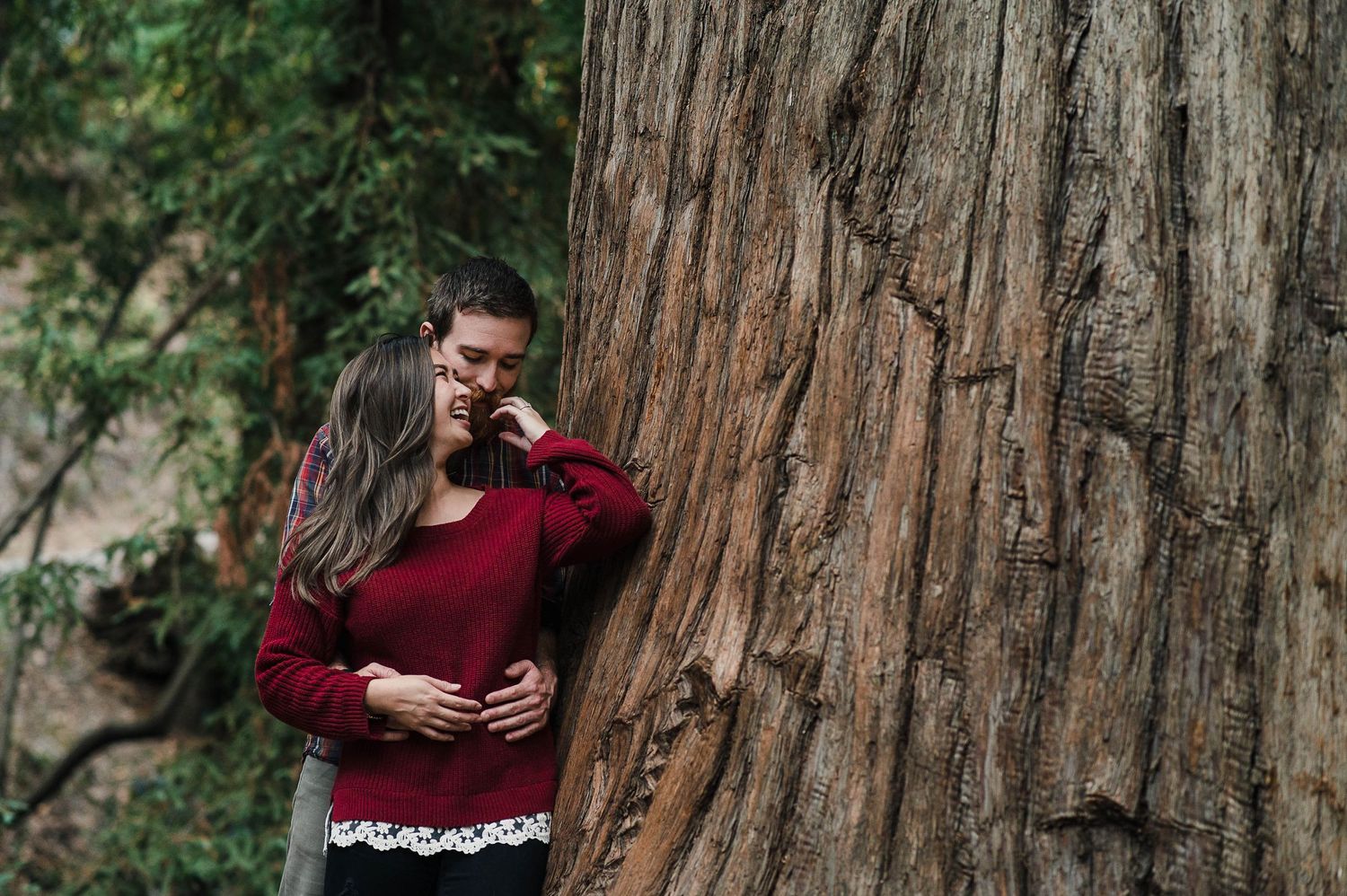 Romantic couple shares intimate moment next to massive redwood tree trunk in forest.