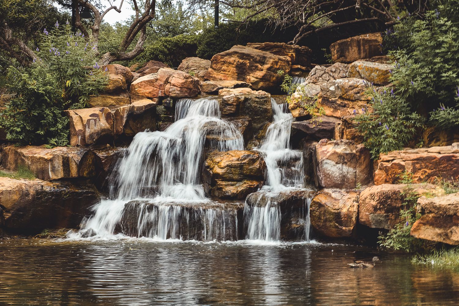 Multi-tiered waterfall cascades over orange rocks into a tranquil pool surrounded by lush vegetation.