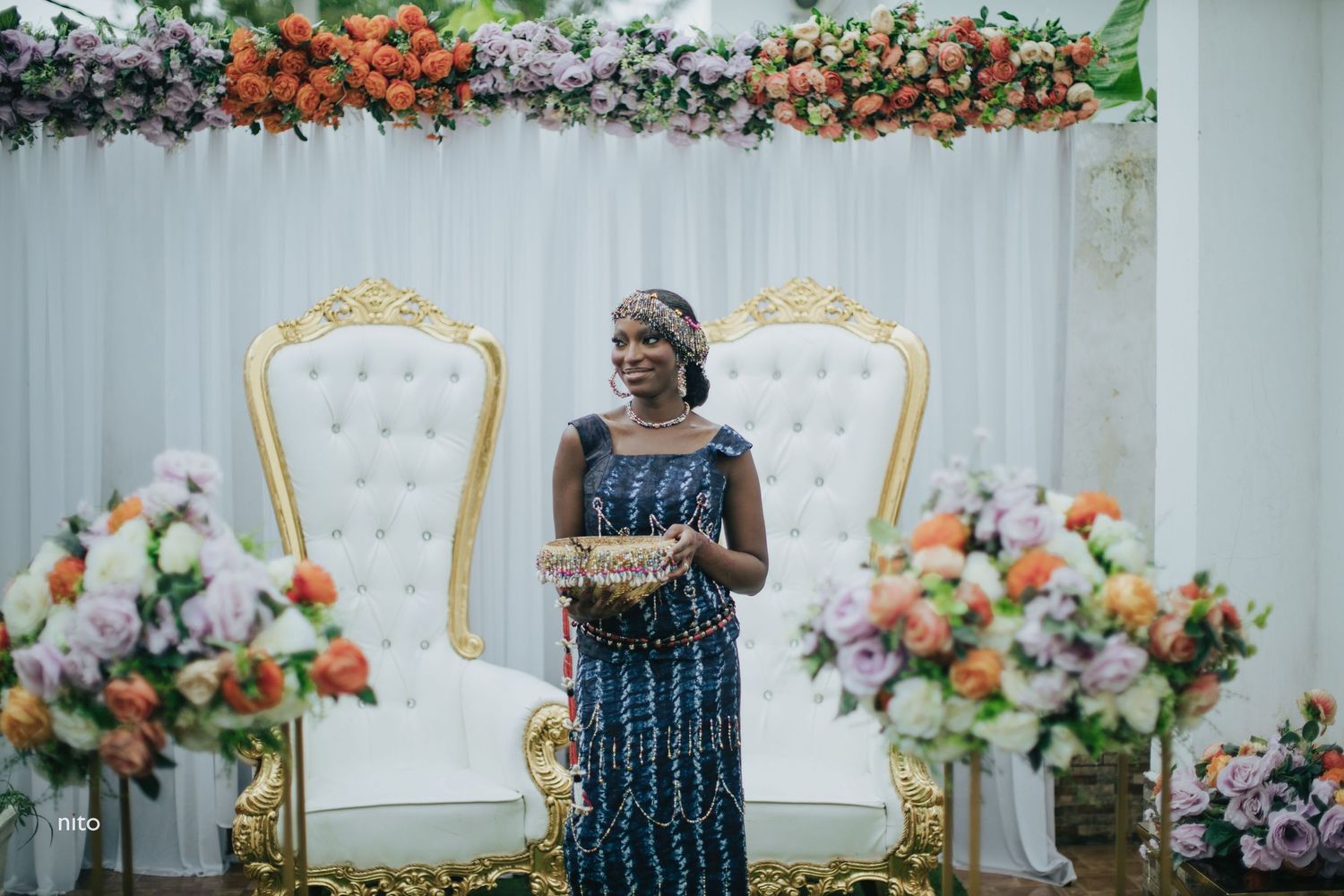 An elegant wedding scene with ornate white chairs and floral arrangements against a decorated backdrop.