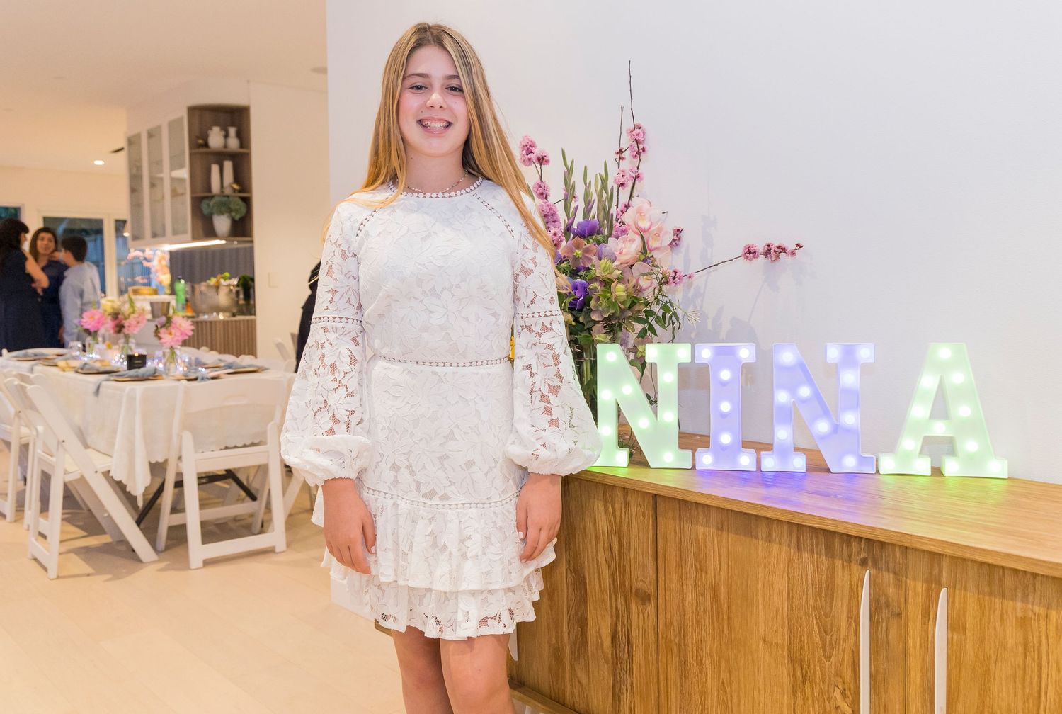 White lace dress celebration event with illuminated NINA sign and floral arrangements.