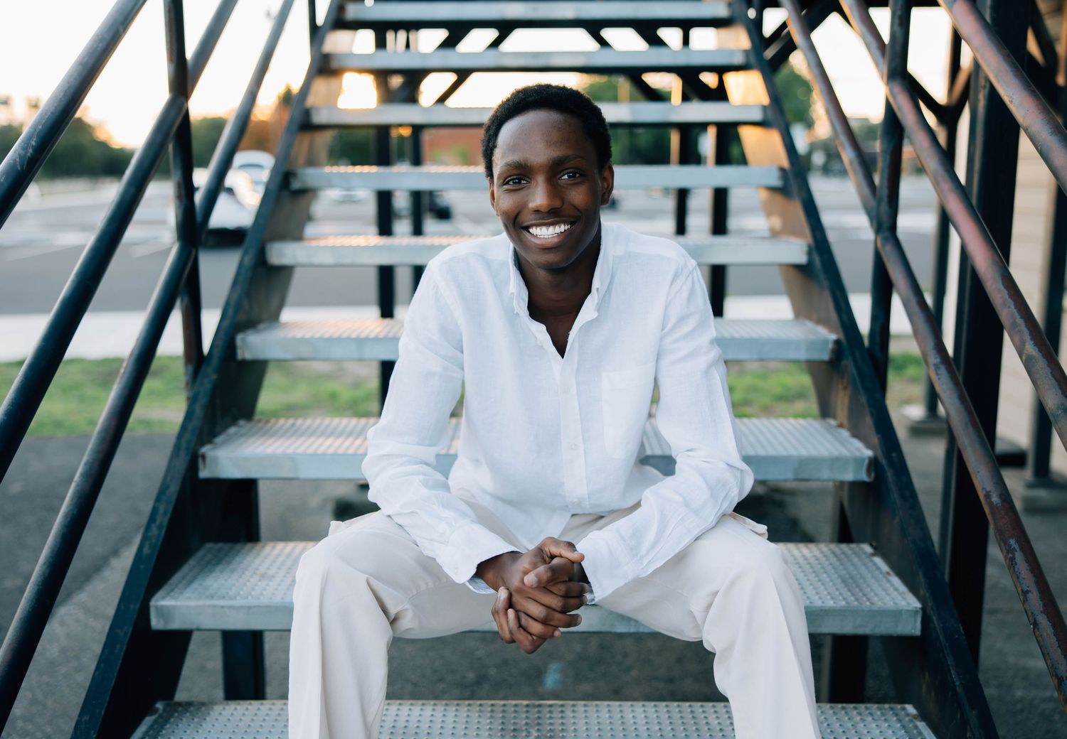 Smiling man in white shirt sits on metal stairs outdoors on a sunny day.