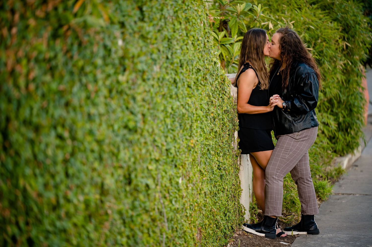 Couple sharing tender moment against ivy-covered wall in natural lighting.