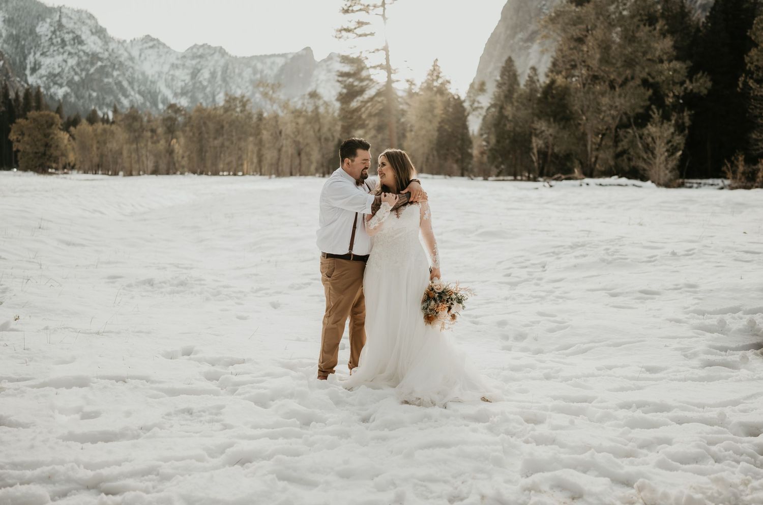 Wedding couple embraces in snowy mountain landscape with evergreen forest backdrop.