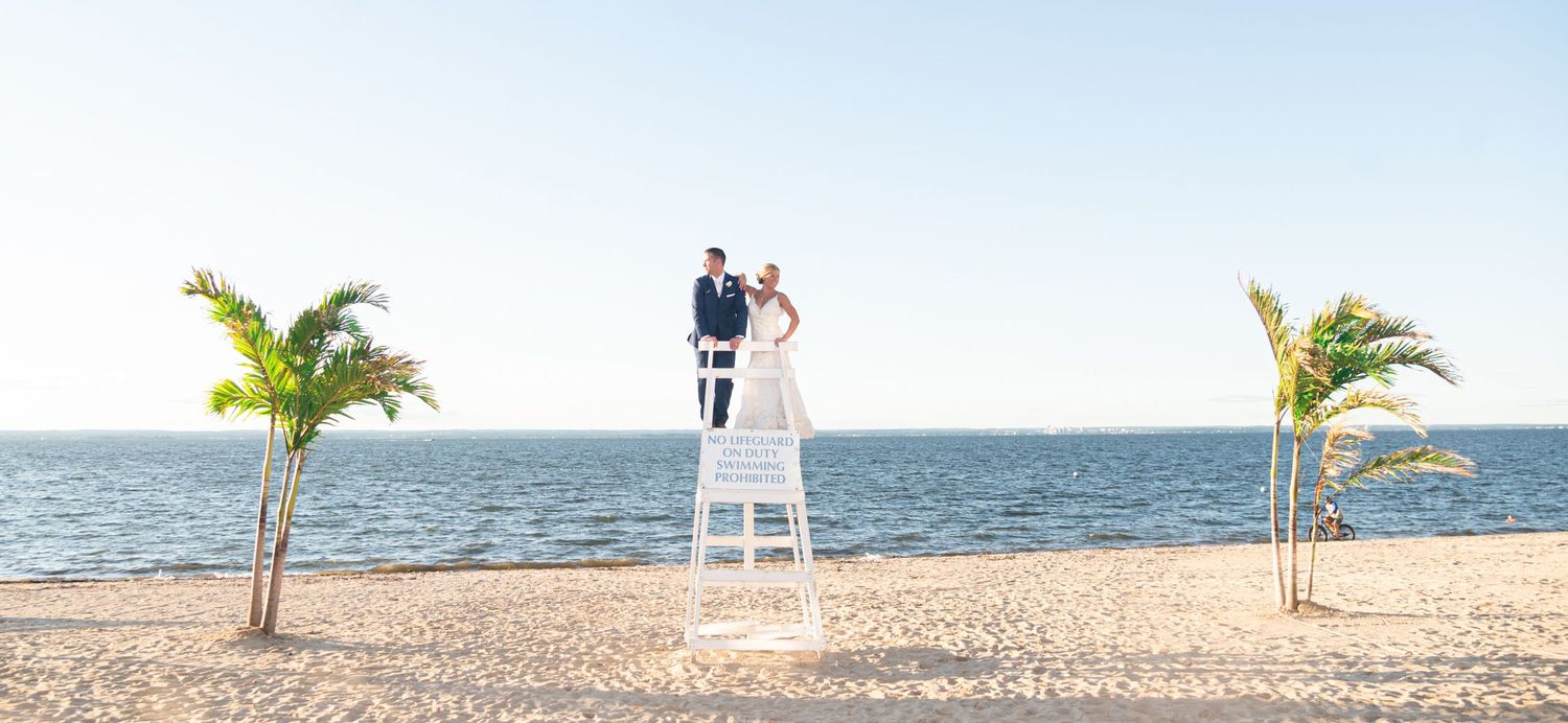 A couple poses standing on the lifeguard chair at Crescent Beach Club before their wedding.