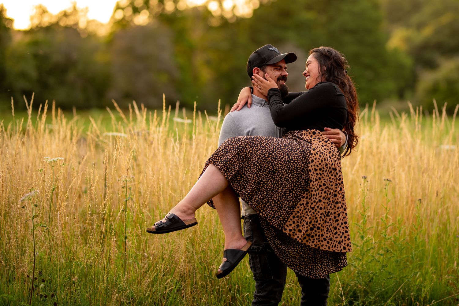 Man lifting fiance off the ground, she holds his face and both are smiling. Bathed in the warm light of a sunset.