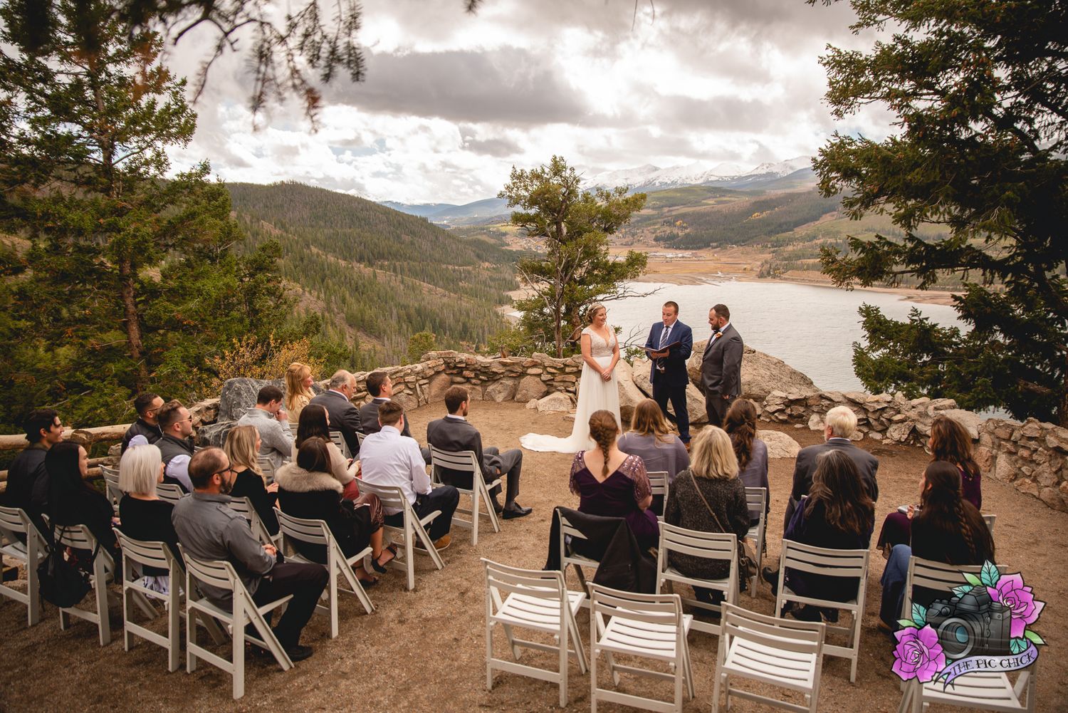 Outdoor mountain wedding ceremony overlooking a scenic lake with guests seated on white chairs.