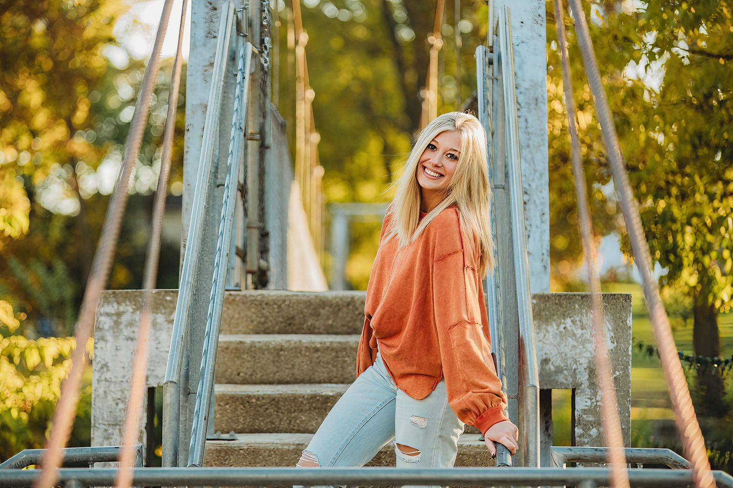 Person in orange top and jeans poses playfully on outdoor steps surrounded by greenery.