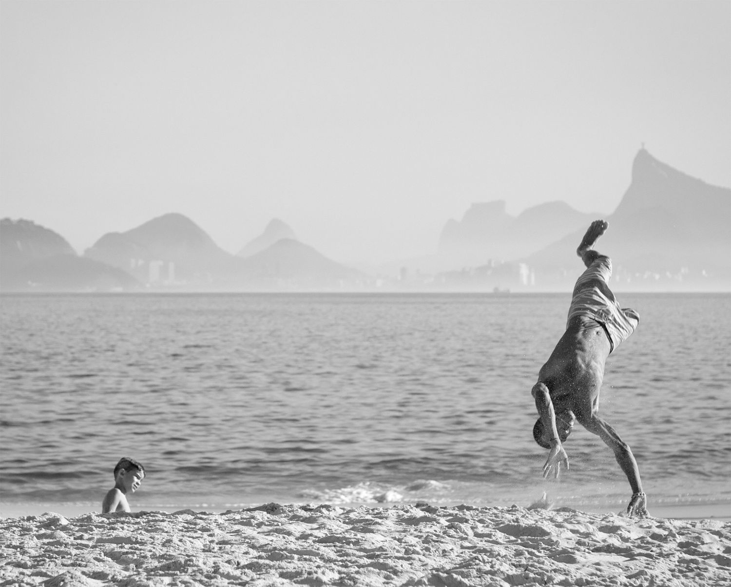 Dog leaping joyfully on a beach with mountains in the background captured in black and white.