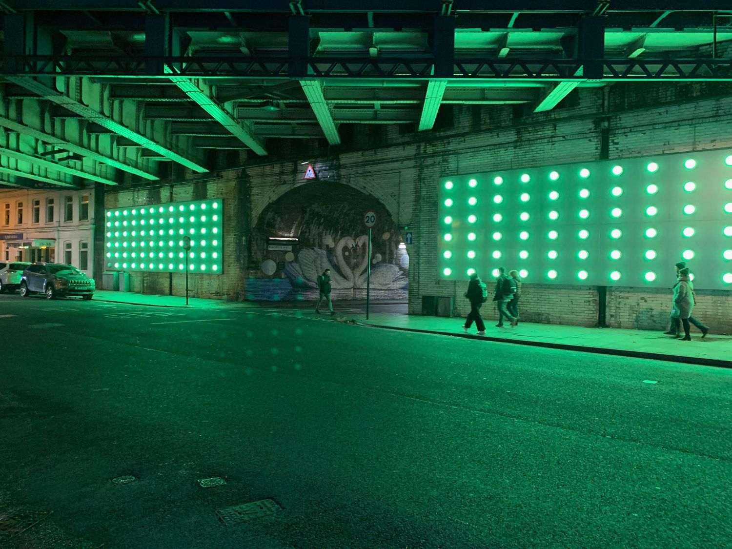 Dramatic green neon light installation under railway arches with graffiti murals in London.