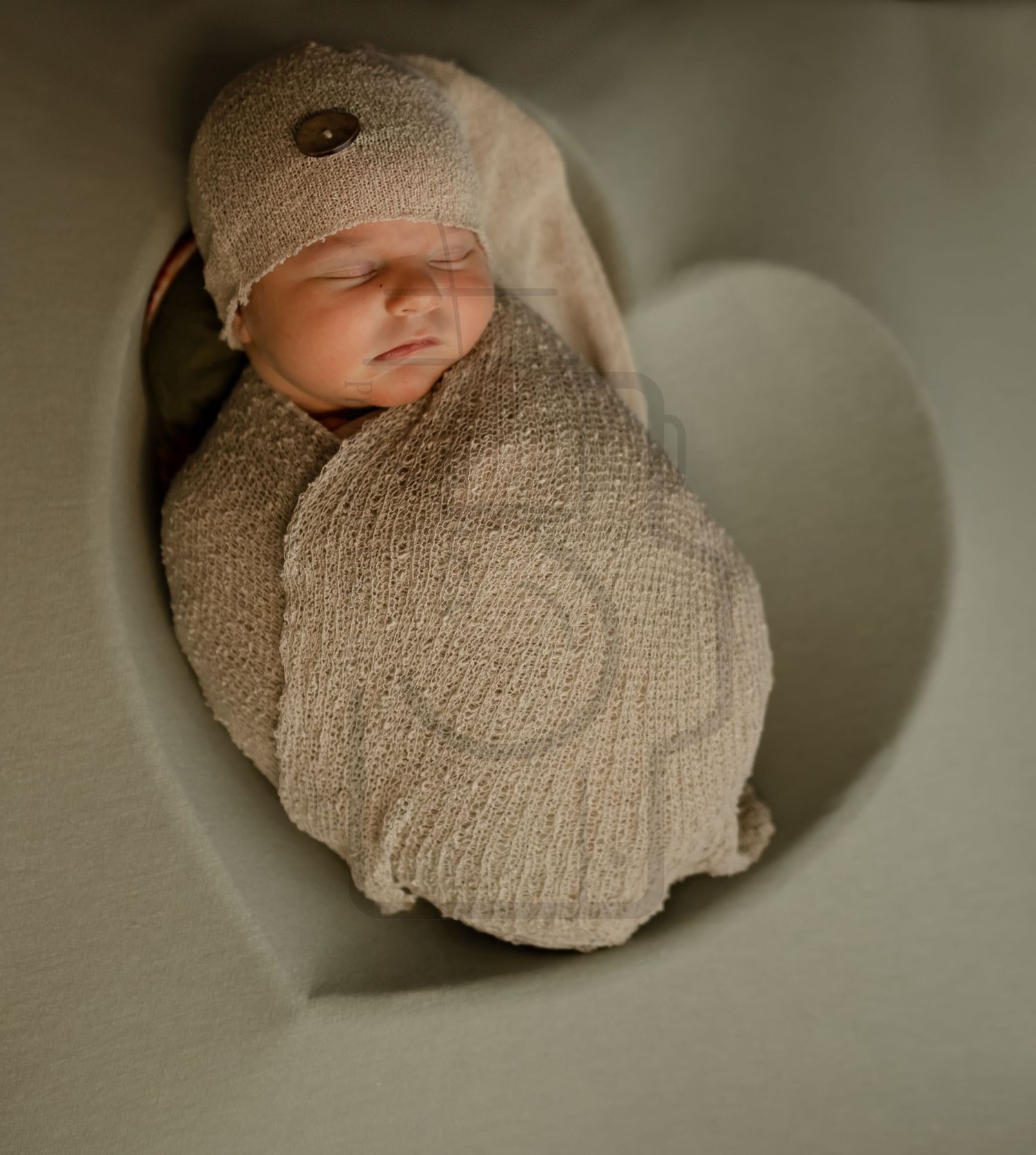 A sleeping newborn wrapped in tan swaddle blanket photographed through a heart-shaped frame.
