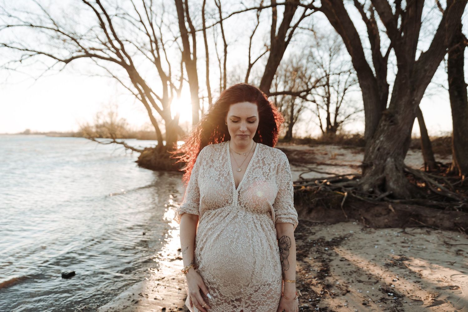 Silhouette of expectant mother in dress standing by water at sunset.