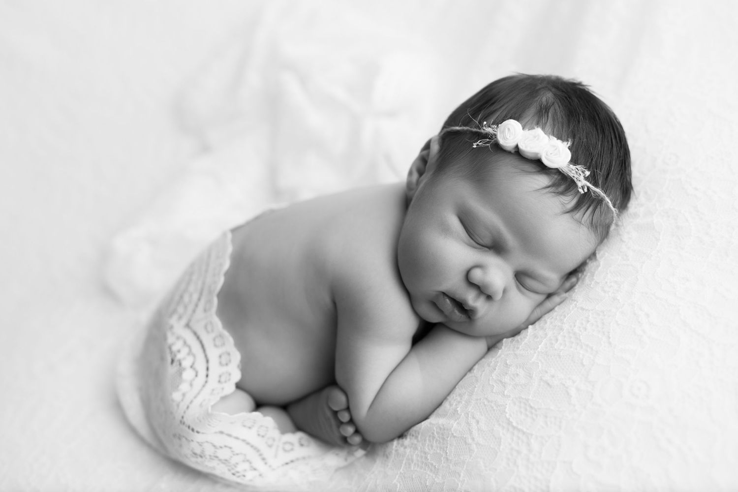 Black and white image of newborn baby girl on white lace blanket, lace wrap around her bottom and floral headband.