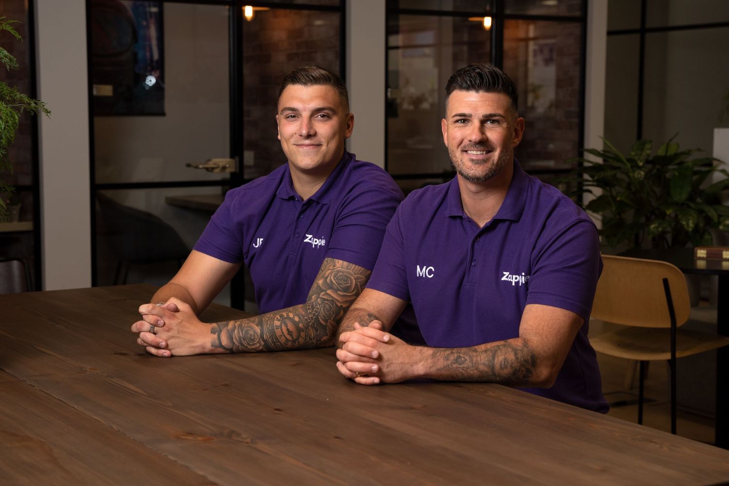 Two colleagues in purple uniforms sitting together at wooden table in office setting.