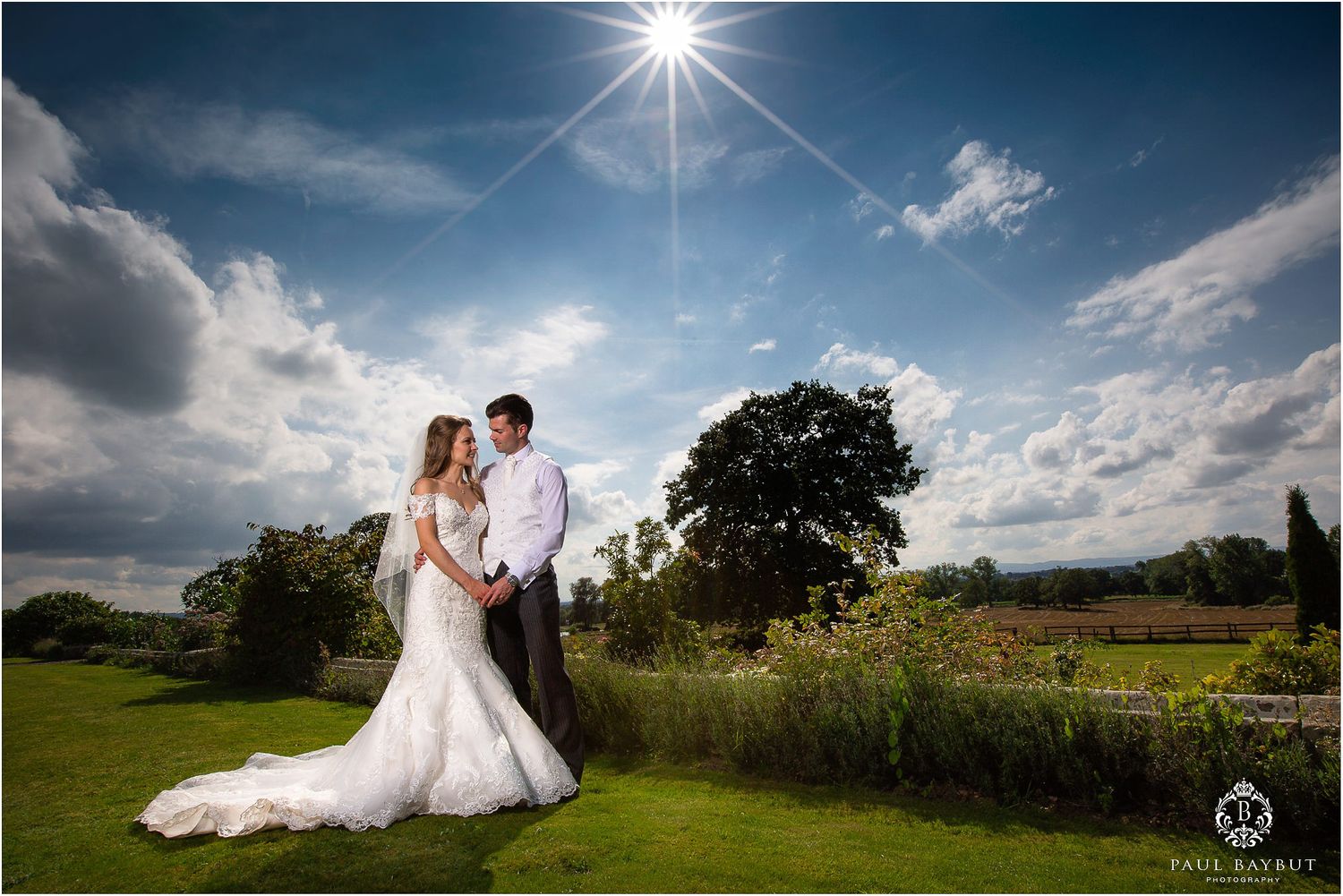 Newlyweds pose in sunlit garden landscape with dramatic sky and sunburst.