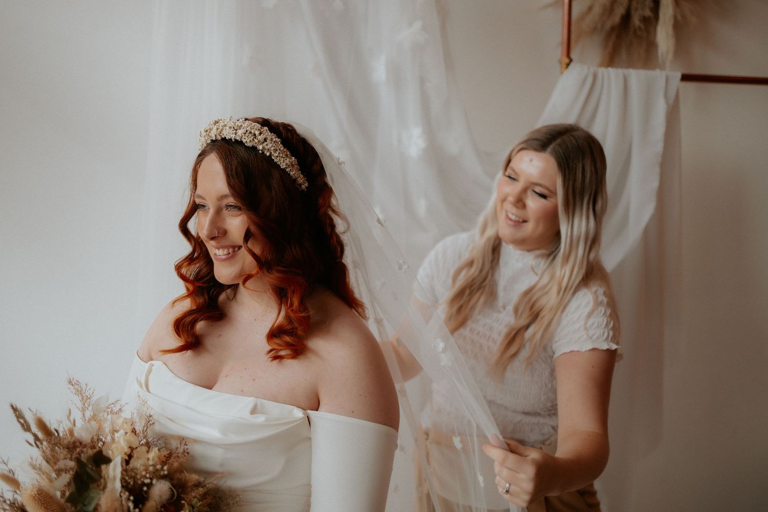A bridal preparation moment captured in soft light with an off-shoulder wedding dress and delicate headpiece.