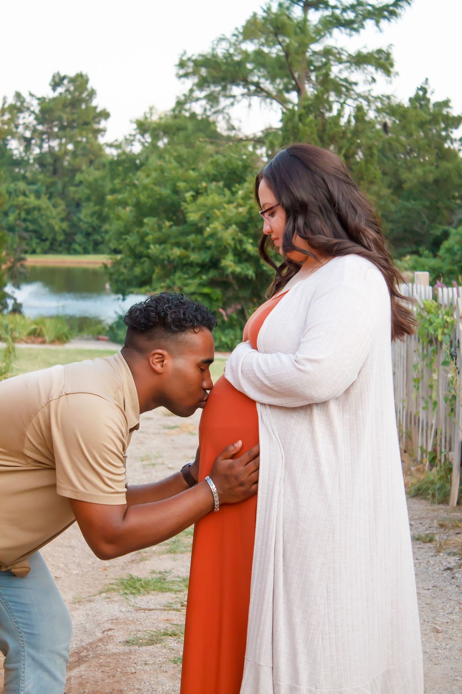 Maternity photo of couple in white and tan outfits by lakeside during golden hour.