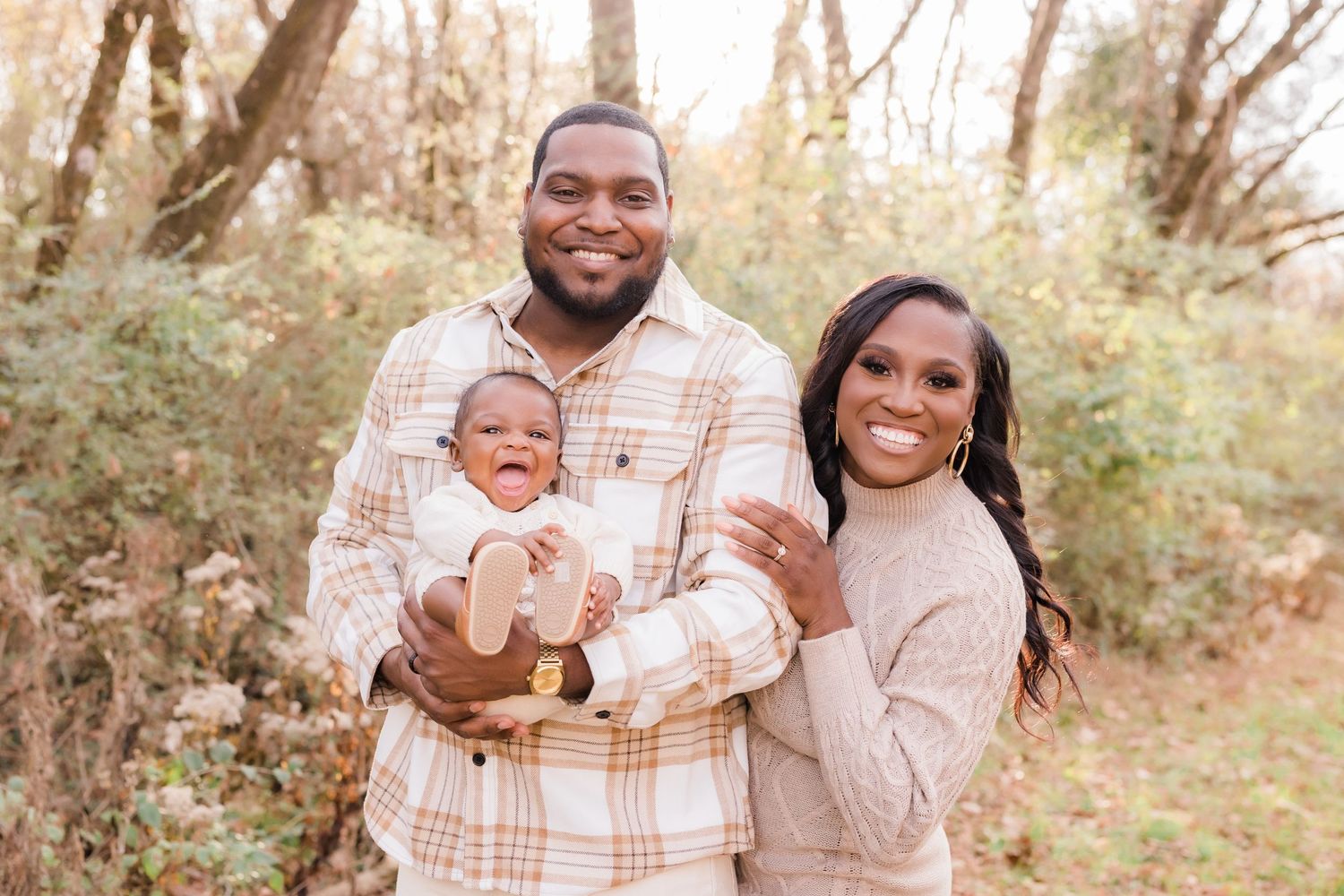 Happy family of three posing outdoors in autumn, parents holding smiling baby in neutral tones.