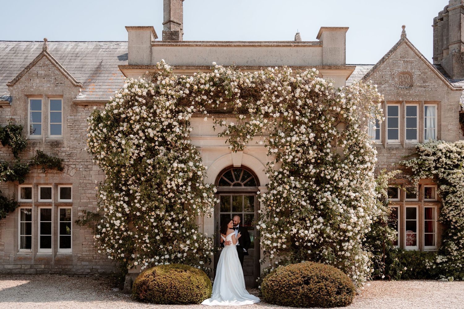 Bride and groom stand together in a doorway surrounded by white flowers at Euridge Manor, Cotswolds Wedding Venue
