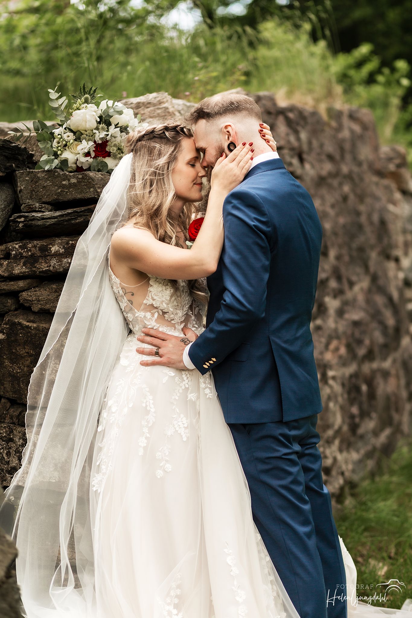 A couple shares an intimate moment against a stone wall with bridal flowers.