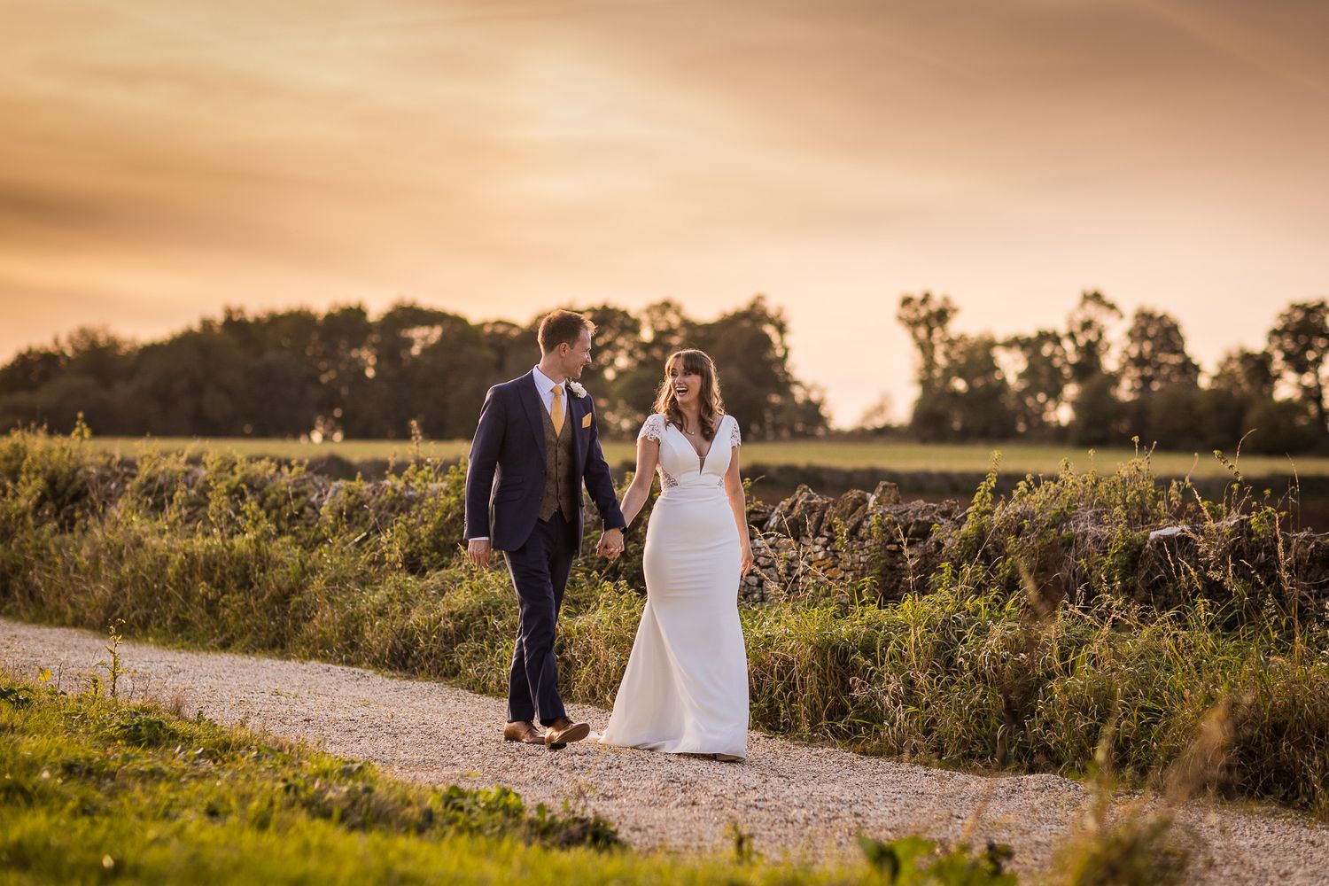 Couple walks along countryside path during golden sunset wedding photo session.