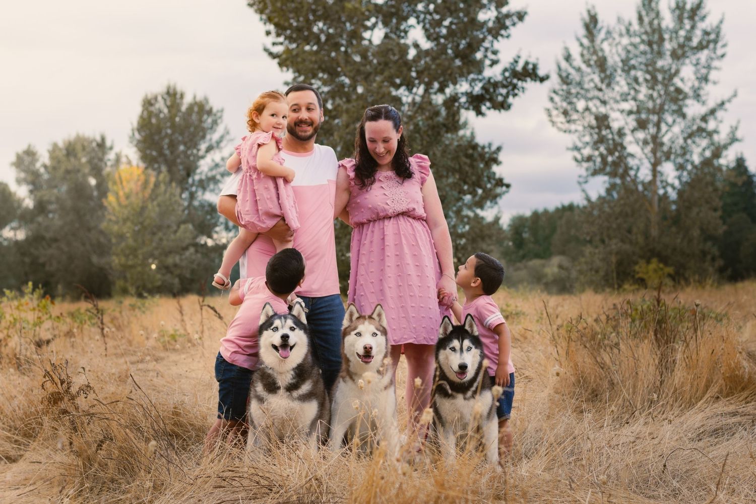 Family posing with in field with trees in background. mom dad 3 kids and 3 huskies. All matching outfits. soft pink.
