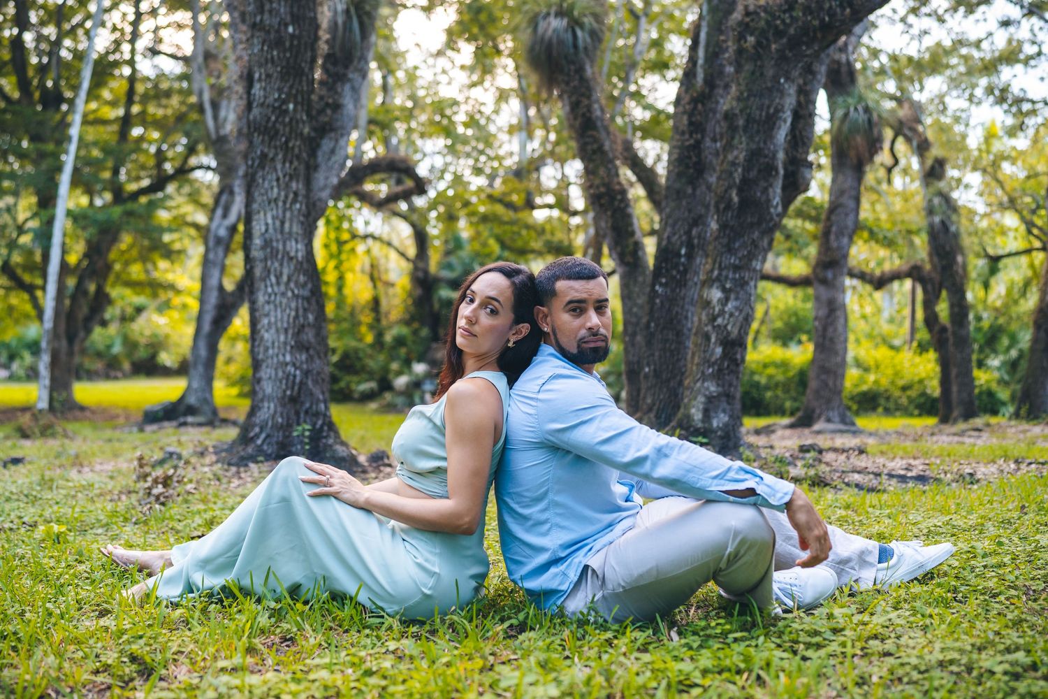 Couple relaxing back-to-back on grass under large trees.