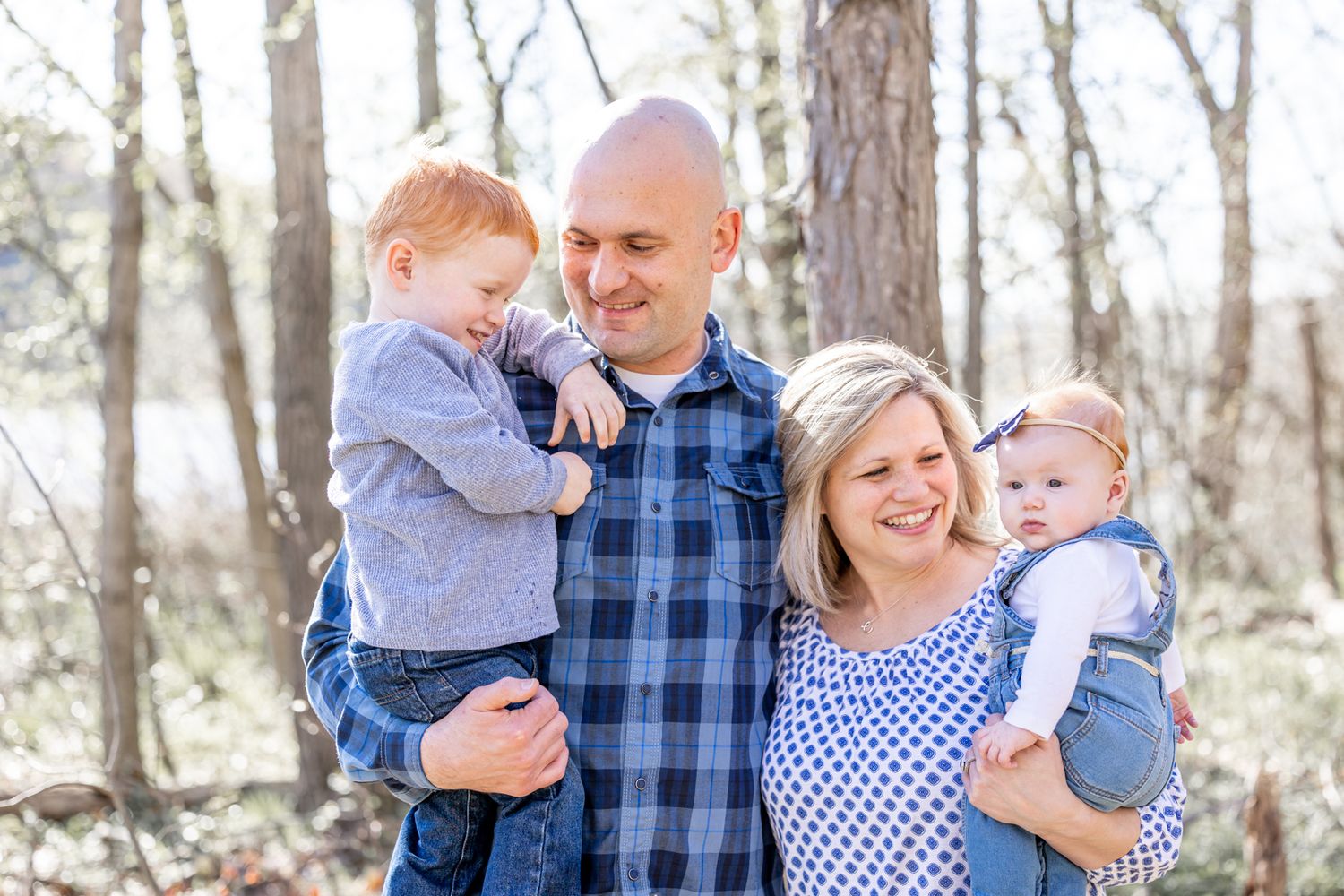 A family in coordinating blue outfits poses together in a wooded outdoor setting during a photo session.