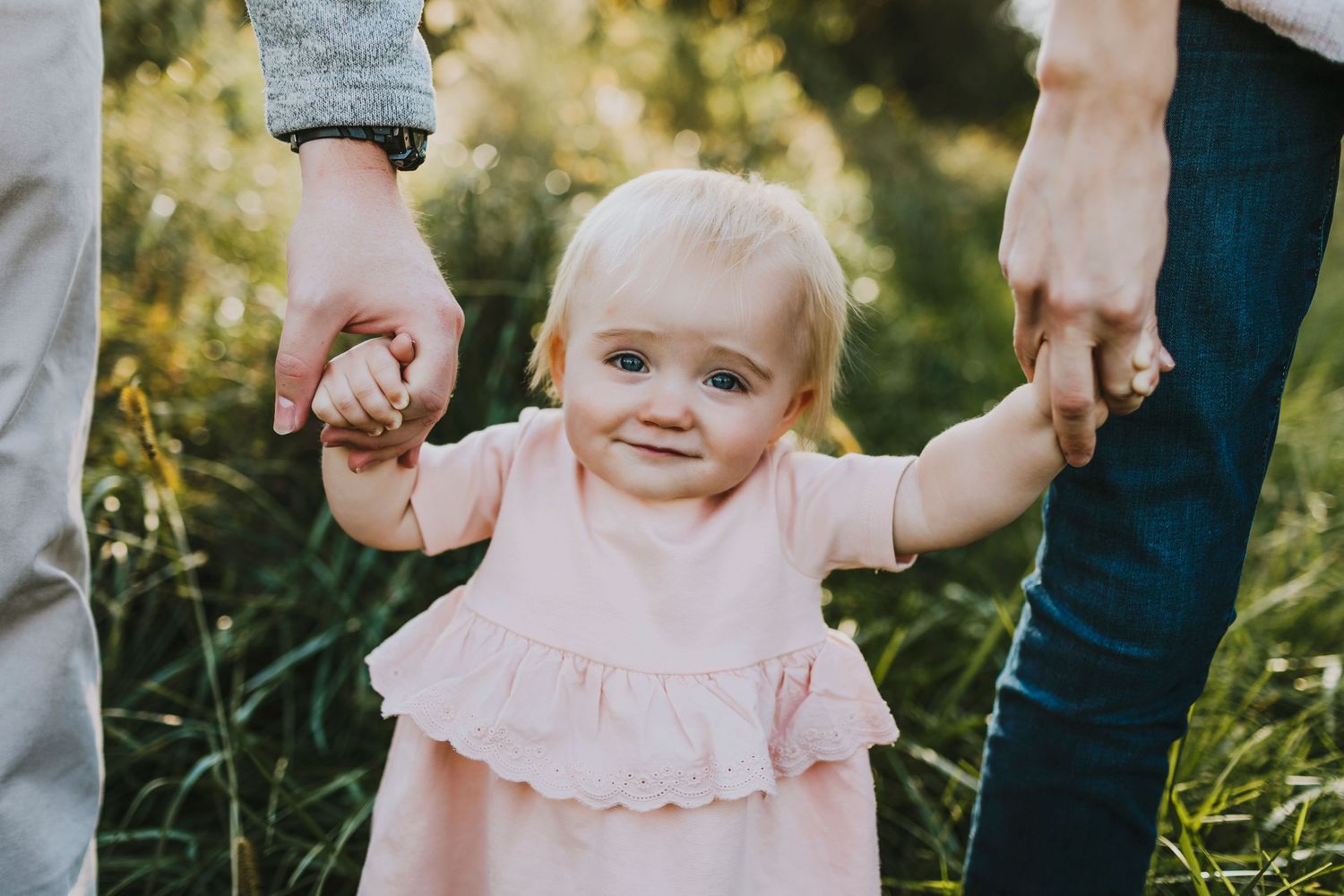 Small child in pink dress holds hands with adults while walking outdoors in sunlit grass.