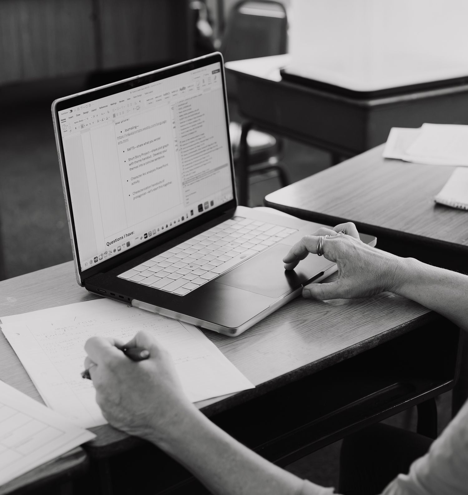 Person working at a wooden desk with an open MacBook laptop and handwritten notes in a black and white photo.